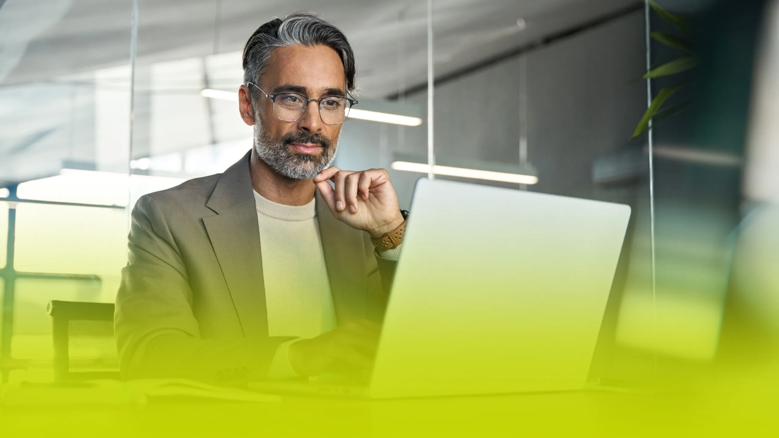 Focused man in a tan blazer using a laptop at a glass-walled office, pendant lights above and a subtle lime gradient in the foreground.